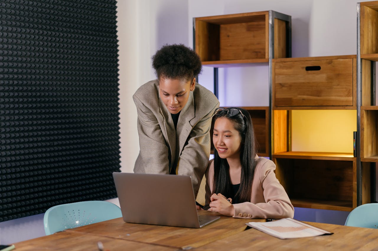 Two women collaborating in a modern office setting, engaging with a laptop and paperwork.